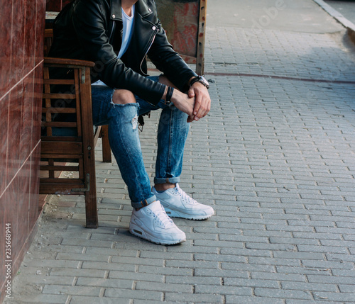 attractive guy sitting on a bench in a cafe, on the street, white sneakers, white T-shirt, black leather jacket and black jeans, pants. model. waiting for a meeting.