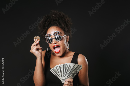 Image of happy afro american woman wearing dollar glasses holding golden bitcoin and money banknotes, isolated over black background