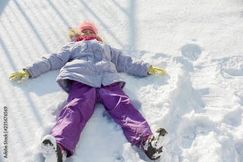 Winter portrait of cheerful child girl having fun in the snow, lying on the snow in the form of angel, top view