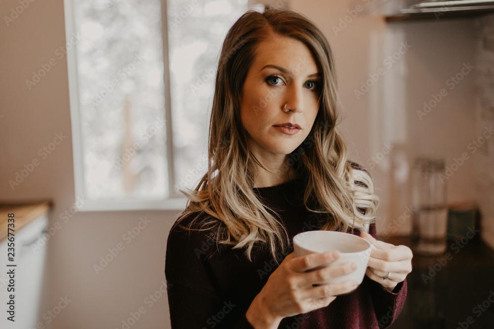 Pretty Young Blonde Woman Sipping Morning Hot Coffee in Modern Designed Kitchen While Looking out Bright Interior Window in the Winter Pretty Young Blonde Woman Sipping Morning Hot Coffee in Modern Designed Kitchen While Looking out Bright Interior Window in the Winter
