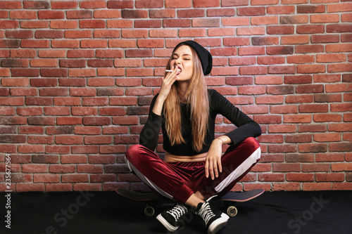 Photo of subcultural sporty girl 20s, sitting on skateboard with cigarette against brick wall photo