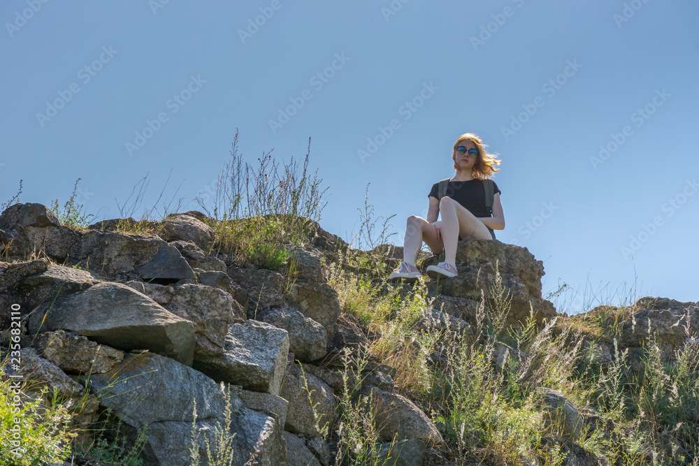 Young pretty tourist lady in sunglasses sitting on stones of ancient architectural attractions ruins on the blue sky background in sunny summer day. Travel concept