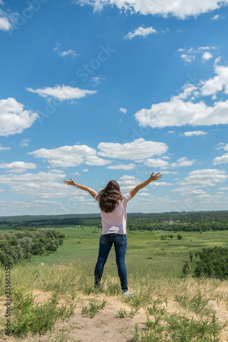 Young hipster girl is travelling in summer holidays. Woman in modern dress is standing on the hill and looking at the field. Blue sky and white clouds. Freedom and lifestyle concept. vertical photo.