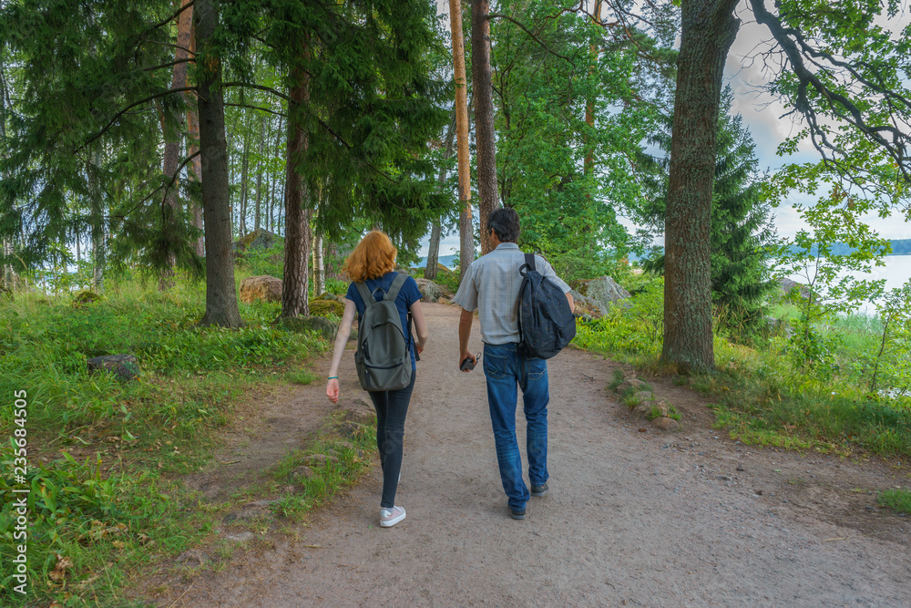 Dark-haired middle-aged man and red-haired young lady walk along forest road. Tourists on the beautiful landscape background. Monrepos Park, Vyborg, Russia. Travel concept