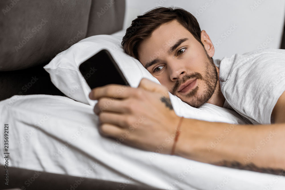 Fototapeta premium handsome bearded young man lying in bed and using smartphone
