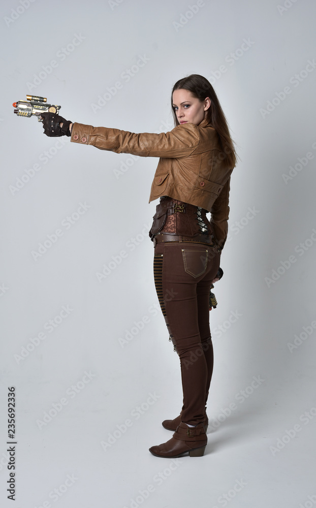 full length portrait of brunette girl wearing brown leather steampunk outfit. standing pose, holding a gun, on grey studio background. full length portrait of brunette girl wearing brown leather steampunk outfit. standing pose, holding a gun, on grey studio background.