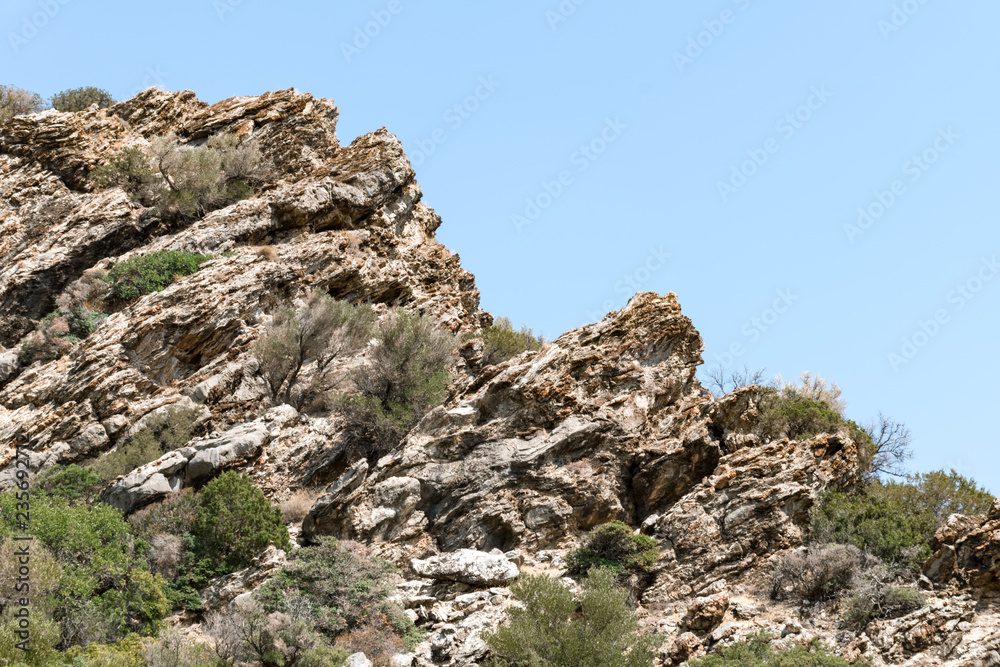 Rocky cliff with rare bushes against light blue sky in Greece.
