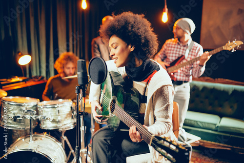 Mixed race woman singing and playing guitar while sitting on chair with legs crossed. In background drummer and bass guitarist. photo