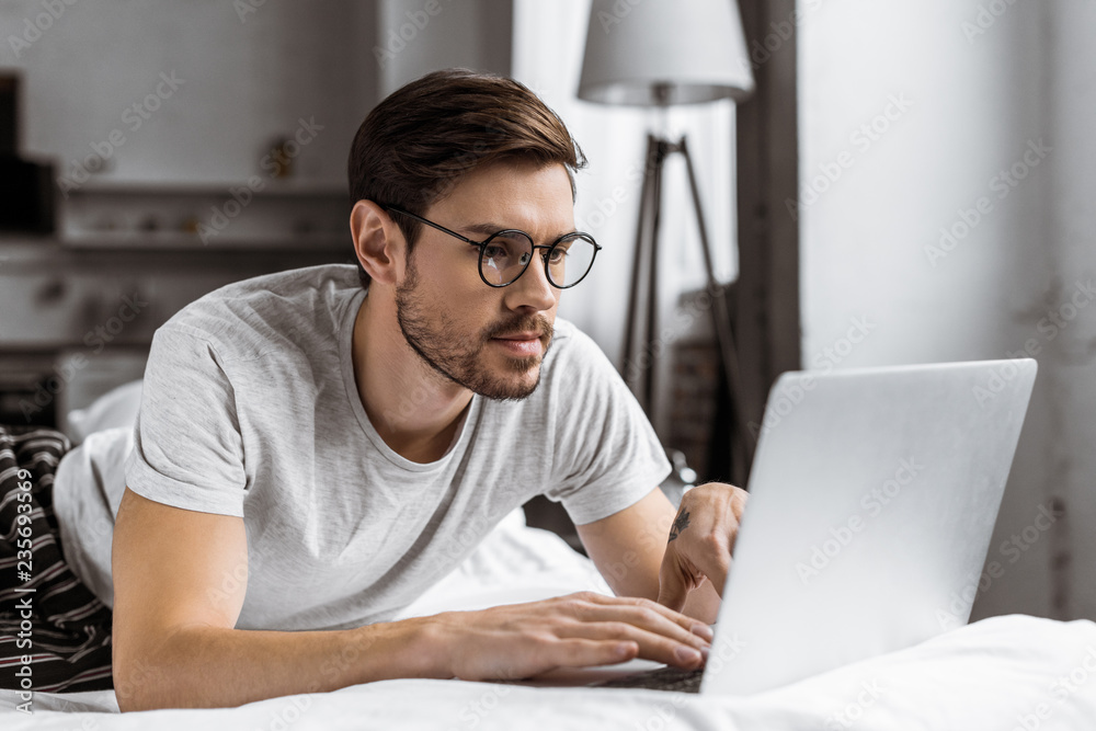 handsome young man in eyeglasses and pajamas using laptop on bed in the morning handsome young man in eyeglasses and pajamas using laptop on bed in the morning