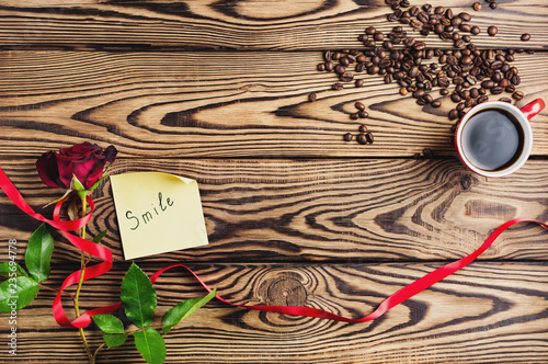 Inscription smile on paper beside red rose with ribbon and cup of coffee near coffee beans on old wooden table. Top view with copy space. Saint Valentine's Day concept