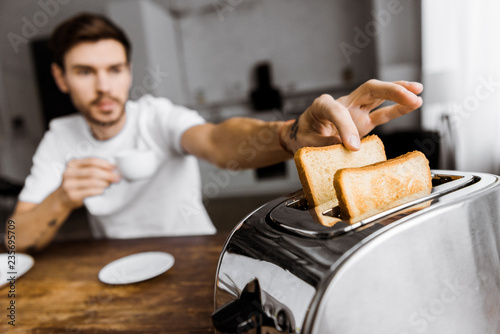 Photos close-up shot of young man drinking coffee and taking toast from toaster