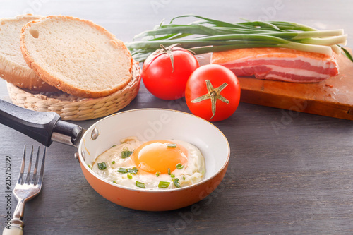 Fried egg in a pan, sliced bread, bacon, tomatoes and green onions cooked for breakfast on a wooden gray table.