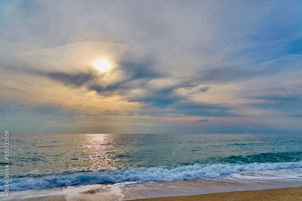                                Sunset at the tropical beach, sun behind clouds reflects on water and waves with foam hitting sand.