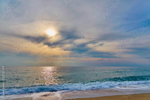  Sunset at the tropical beach, sun behind clouds reflects on water and waves with foam hitting sand.