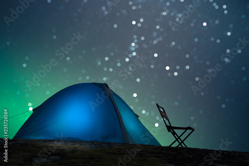 Empty camping chair. Tent with lighting and starry bokeh in the background. Slight aurora. Focus on the chair.