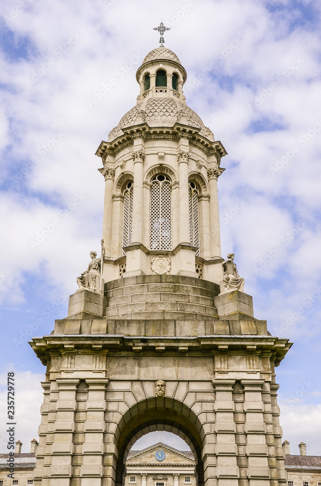 Ornate bell tower under blue sky and puffy white clouds - The Trinity College Campanile in Dublin, Ireland with the Regent House roof and clock centered in the arch of the tower