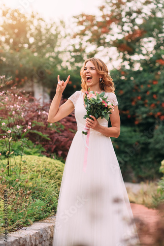 young blonde girl in an elegant long white light dress is joking laughing at the camera in a green garden, holding a lovely bouquet of pink flowers. happy wedding day of the bride. emotional photo