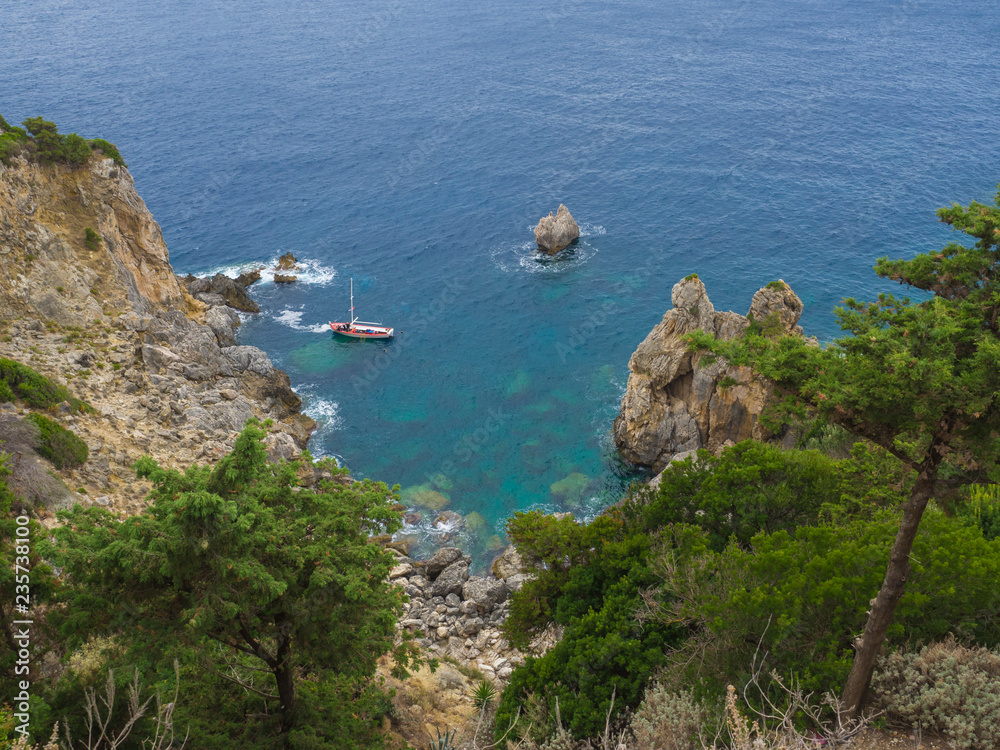 View on cllifs, trees and green hill at Paleokastritsa bay with small red boat, summer cloudy sky, Corfu, Kerkyra, Greece