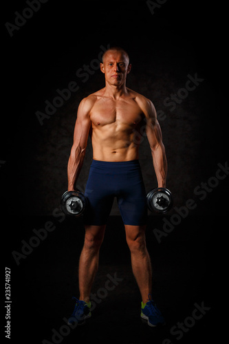 Handsome muscular man working out with dumbbells over black background.