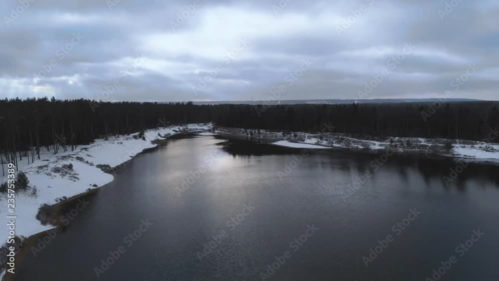 Aerial view. The lake in the winter forest. Flying over a not frozen lake in cloudy winter weather. Colorful landscape, frosty evening. Winter Russian nature. Lake surrounded by snow. 4K Aerial view. The lake in the winter forest. Flying over a not frozen lake in cloudy winter weather. Colorful landscape, frosty evening. Winter Russian nature. Lake surrounded by snow. 4K