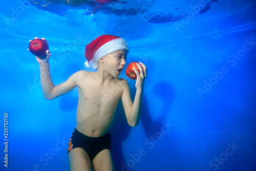 The child prepared to bite off the red Apple, which he holds in his hand. He poses underwater on a blue background in Santa's hat. Concept. Close up. Horizontal orientation