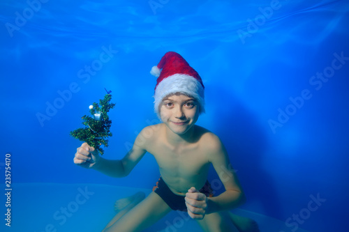 A child with a small Christmas tree in his hand posing and smiling under the water on a blue background in the hat of Santa Claus. Portrait. Close up. Horizontal orientation