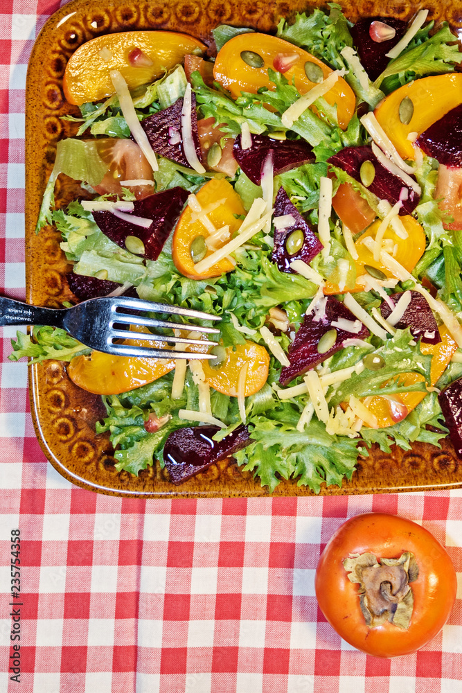 Fototapeta premium close - up of a delicious autumn salad with chocolate persimmon, lettuce, tomatoes, pomegranate on a clay plate with ingredients on a napkin with a red cage, top view