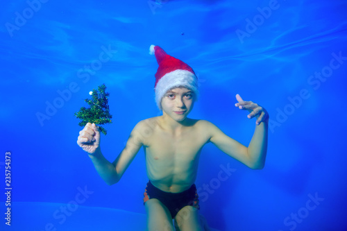 A little boy in a Santa Claus hat with a Christmas tree in his hand swims and poses under the water on a blue background and looks at the camera. Portrait. Concept. Horizontal orientation