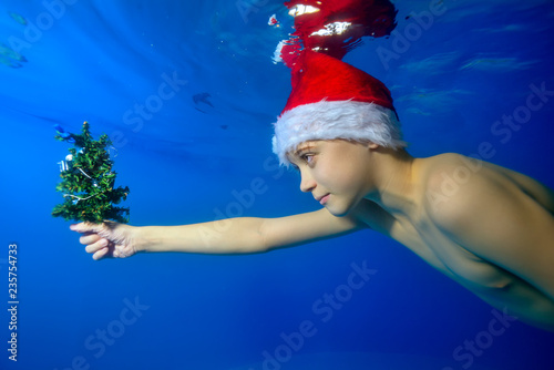 Portrait of a child who swims underwater in the pool with an outstretched hand in which holds a small Christmas tree. He's wearing a Santa hat. Portrait. Side view. Horizontal orientation