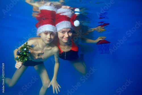 Happy mom and little son swim together underwater in the pool in Santa's hats, holding Christmas toys in their hands. They look at the camera and smile. Landscape orientation