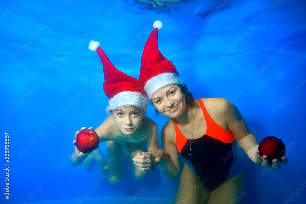 Mom and little boy in Santa's hats smile and pose together under the water on a blue background, holding apples in his hands and looking at the camera. Portrait. Concept. Landscape orientation Mom and little boy in Santa's hats smile and pose together under the water on a blue background, holding apples in his hands and looking at the camera. Portrait. Concept. Landscape orientation