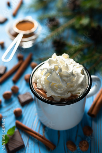 Mug with hot chocolate and whipped cream among cinnamon sticks, nuts on a blue wooden table