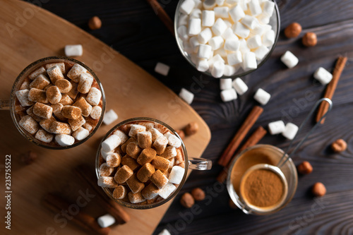 Cups with hot chocolate and marshmallows on cutting board between cinnamon, top view