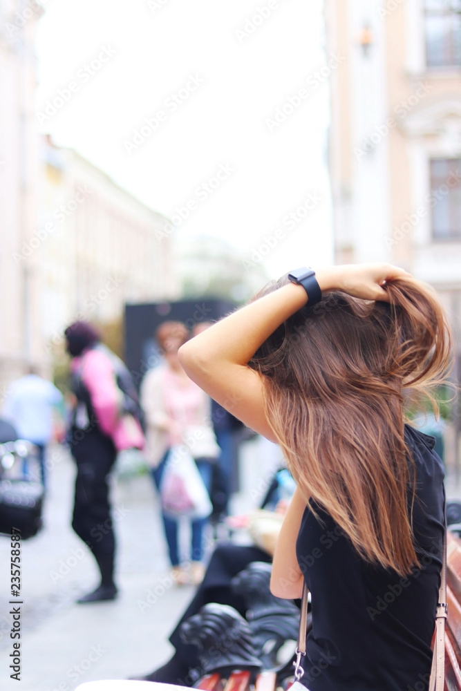 A young girl straightens her long hair while sitting on a bench. A woman sits on a bench in the center of an ancient city against a background of a blurred motorcycle and passers-by. Lifestyle and lei