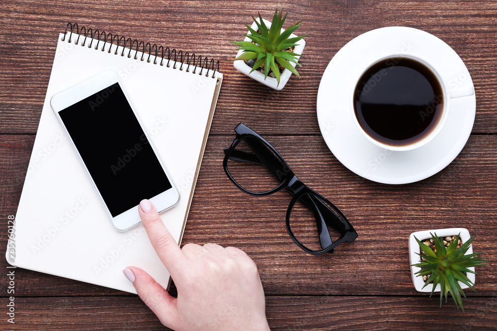 Female finger pressing smartphone button with notebook, cup of coffee and green plants on wooden table Female finger pressing smartphone button with notebook, cup of coffee and green plants on wooden table