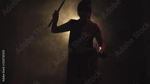 interesting video, a samurai brunette girl with a long pony tail and with two katanas in her hands is standing in a dark room with a dim light, posing and slowly turning to the camera photo