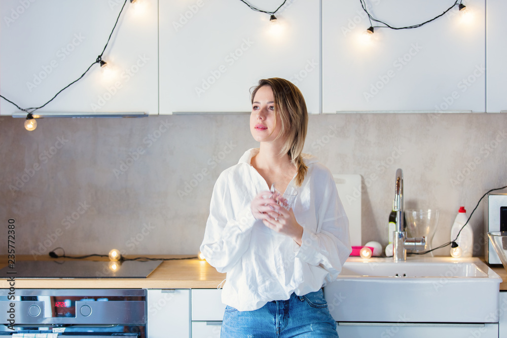 Young caucasian woman in white shirt and blue jeans with glass of a water stay in a kitchen. Young caucasian woman in white shirt and blue jeans with glass of a water stay in a kitchen.