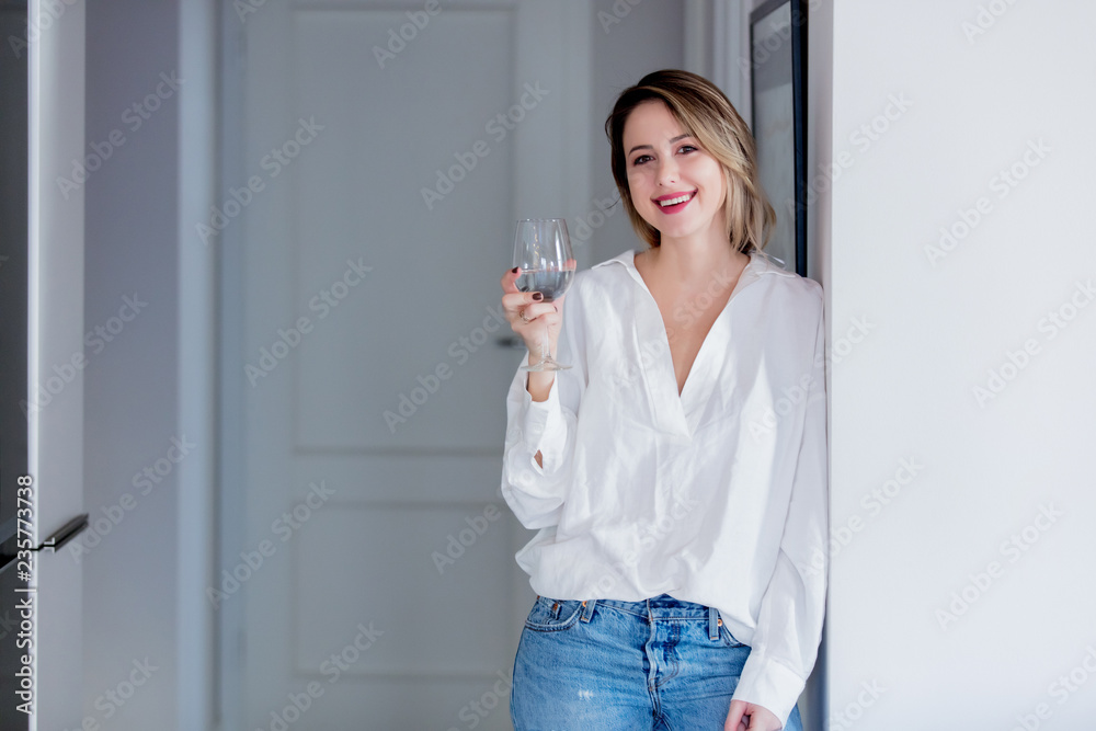 Young caucasian woman in white shirt and blue jeans next to wall with glass of wine. Young caucasian woman in white shirt and blue jeans next to wall with glass of wine.