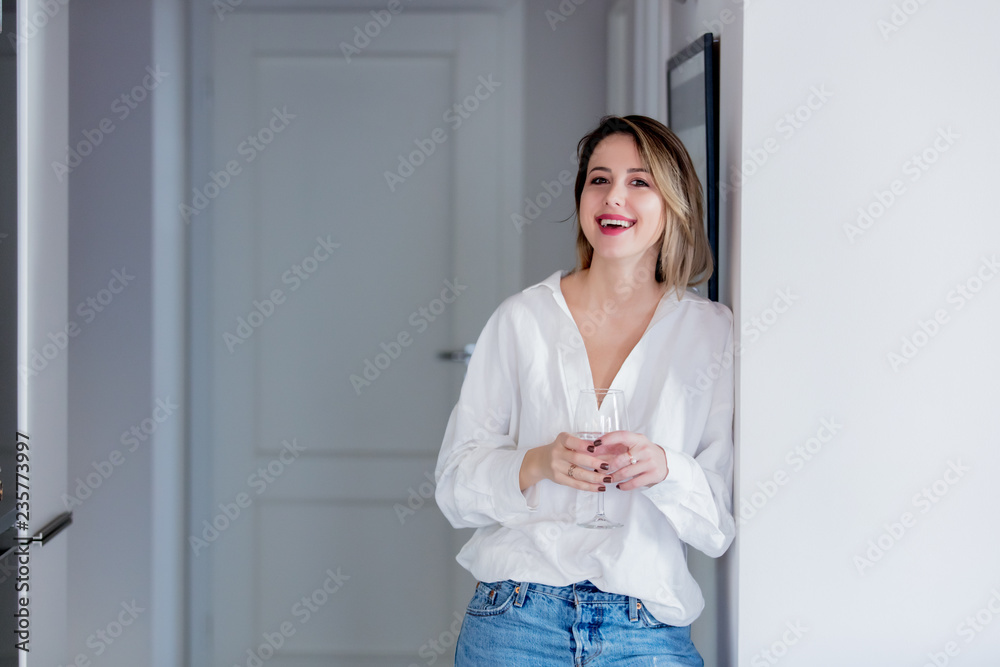 Young caucasian woman in white shirt and blue jeans next to wall with glass of wine. Young caucasian woman in white shirt and blue jeans next to wall with glass of wine.