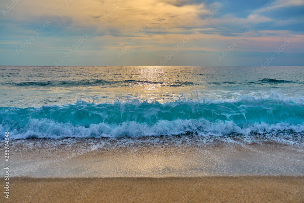 Sunset at the tropical beach, sun behind clouds reflects on water and waves with foam hitting sand.