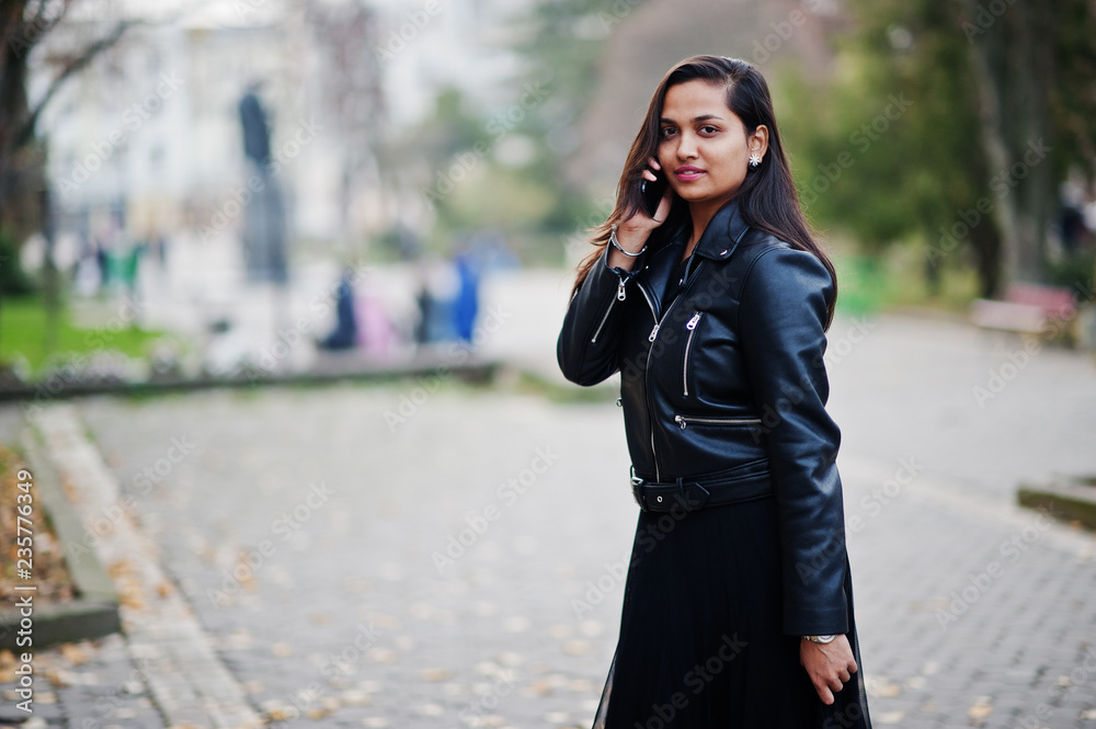 Pretty indian girl in black saree dress and leather jacket posed outdoor at autumn street and speaking on mobile phone.