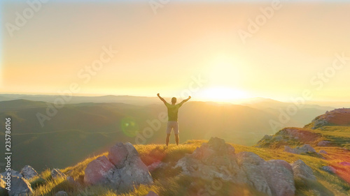 AERIAL: Cheerful man outstretches arms victoriously while observing the sunset.