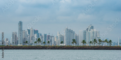 Hot, humid day in Panama city as another rainstorm brews quickly over the city skyline. Tall buildings shimmer in heatwaves rising in humid air. People on Panama Canal jetty park in foreground.