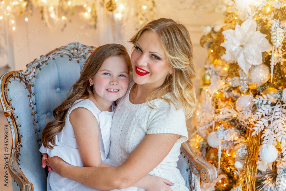 Happy family woman mother and little girl relax playing near Christmas tree on Christmas eve at home. Mom, daughter in light bedroom with winter decoration. Christmas New Year time for celebration Happy family woman mother and little girl relax playing near Christmas tree on Christmas eve at home. Mom, daughter in light bedroom with winter decoration. Christmas New Year time for celebration