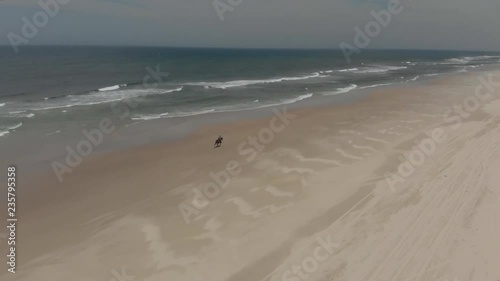 Ascending aerial following horse rider on sandy beach. Blue sky & ocean waves.