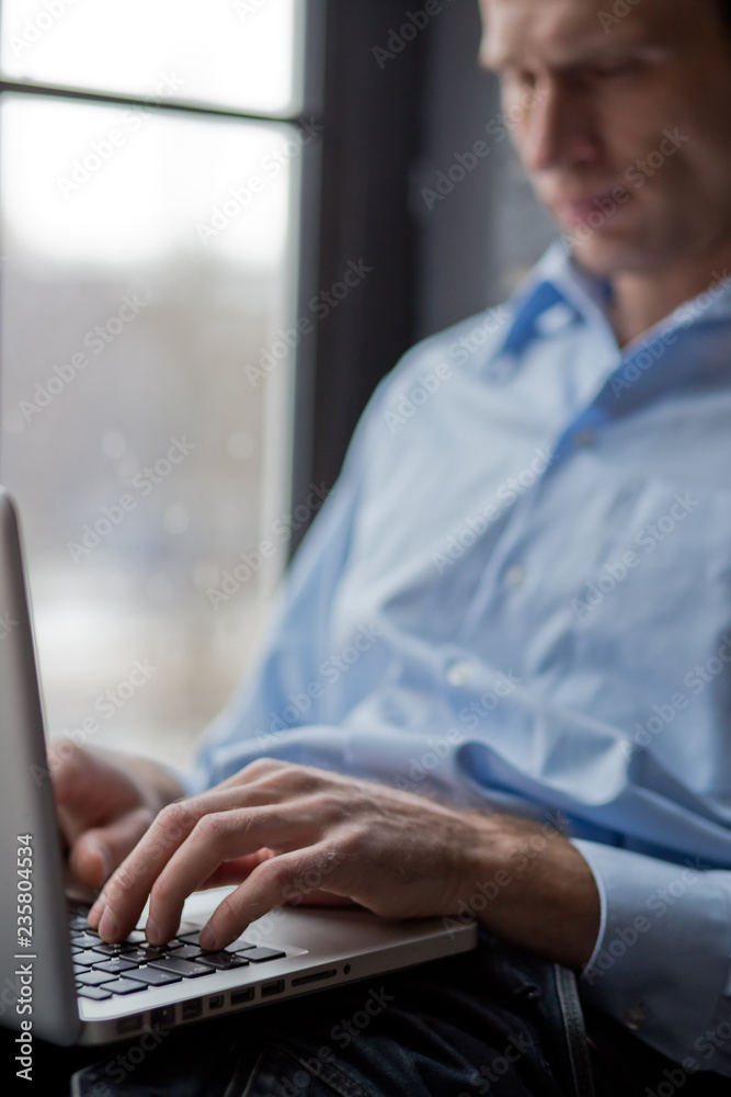 A man sits on a windowsill and works on a laptop. Beautiful man's wrist. Long fingers of a young man. A man sits on a windowsill and works on a laptop. Beautiful man's wrist. Long fingers of a young man.