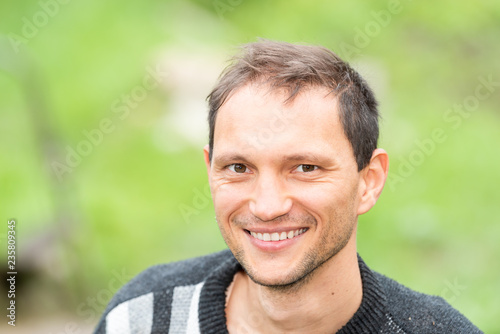 Young handsome attractive happy man face closeup with stubble, showing teeth farmer in garden green summer background portrait