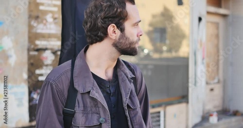 Bearded man with guitar strapped to his back turns to look at woman and window reflection of woman walking away towards the bus