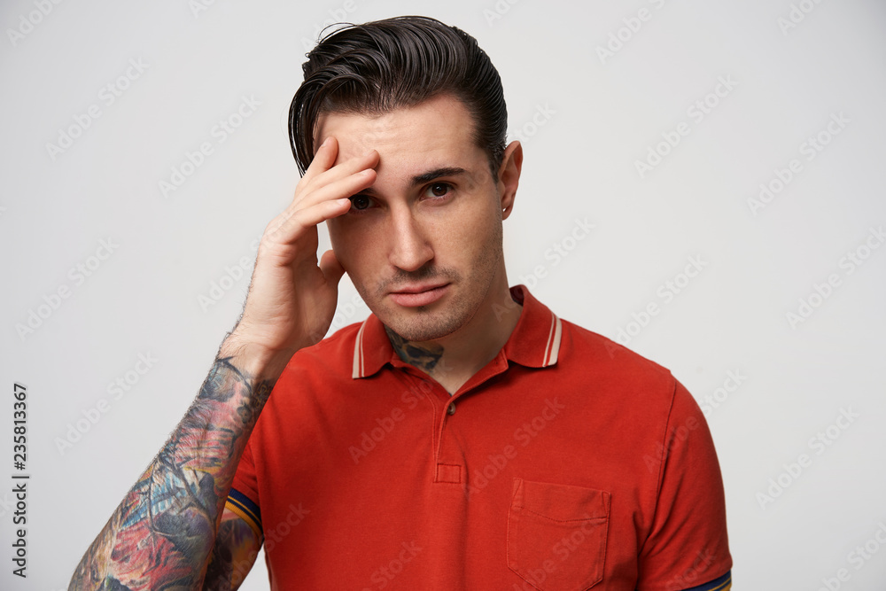 Handsome young man looking at camera and keeping hand near the face while standing against white background. Portrait of attractive self-assured guy with stylish haircut wearing red polo, macho Handsome young man looking at camera and keeping hand near the face while standing against white background. Portrait of attractive self-assured guy with stylish haircut wearing red polo, macho