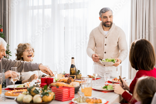 Warm-toned portrait of big happy family enjoying Christmas dinner together, focus on smiling man serving food, copy space